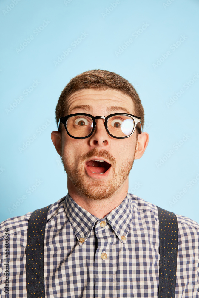 Portrait of young man in checkered shirt, shorts and suspenders with laptop isolated over blue background. Excited and shocked.