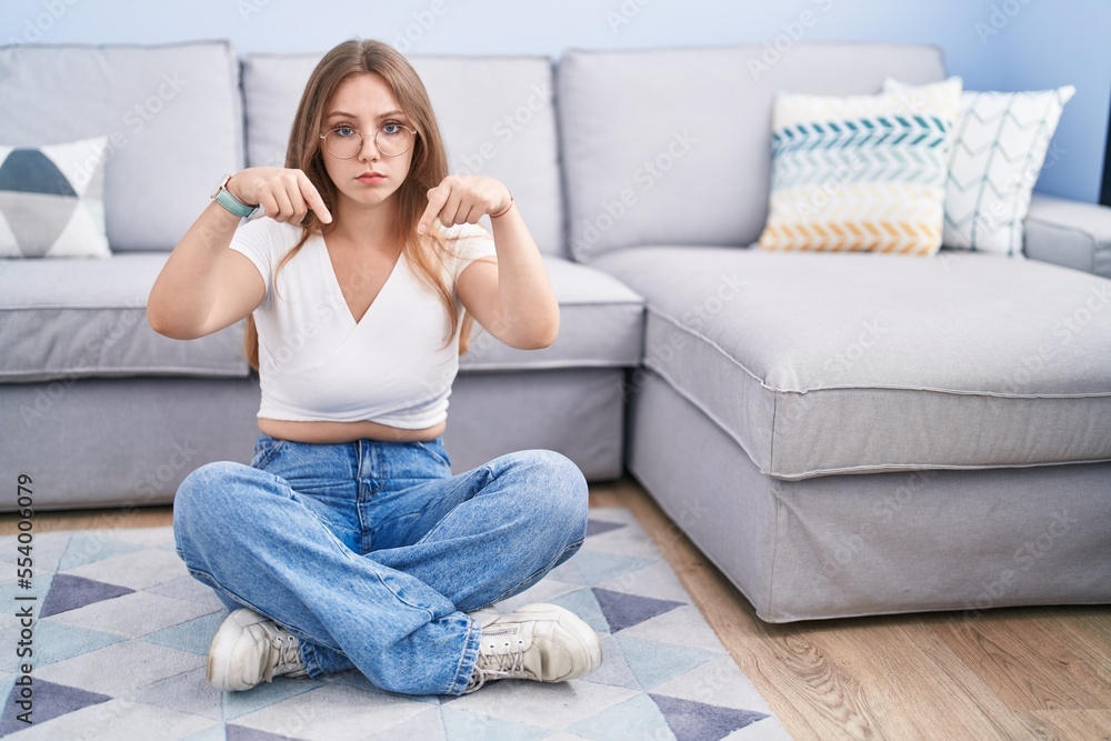 Young caucasian woman sitting on the floor at the living room pointing down looking sad and upset, indicating direction with fingers, unhappy and depressed. Young caucasian woman sitting on the floor at the living room pointing down looking sad and upset, indicating direction with fingers, unhappy and depressed.