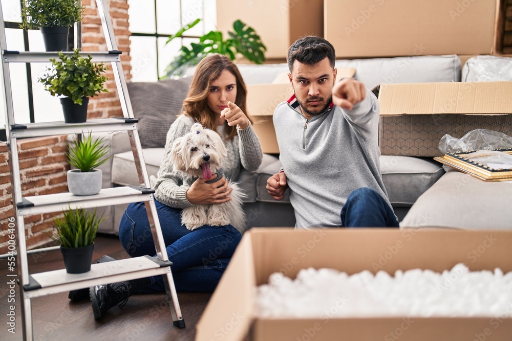 Young hispanic couple sitting on the floor at new home with log pointing with finger to the camera and to you, confident gesture looking serious Young hispanic couple sitting on the floor at new home with log pointing with finger to the camera and to you, confident gesture looking serious