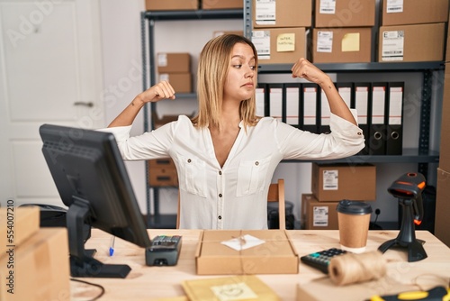 Young blonde woman working at small business ecommerce showing arms muscles smiling proud. fitness concept.