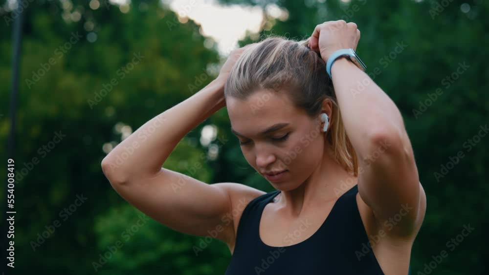 Caucasian athlete woman ties her ponytail before the training, wearing smartwatch tracking her calories on the park background. People and sport concept. Caucasian athlete woman ties her ponytail before the training, wearing smartwatch tracking her calories on the park background. People and sport concept.