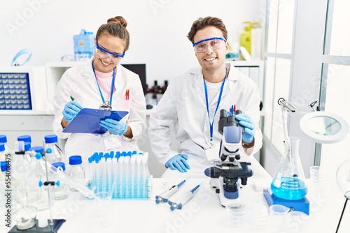 Man and woman wearing scientist uniform using microscope write on clipboard at laboratory