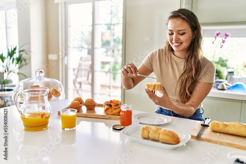 Young beautiful hispanic woman preparing breakfast putting jam on bread at the kitchen