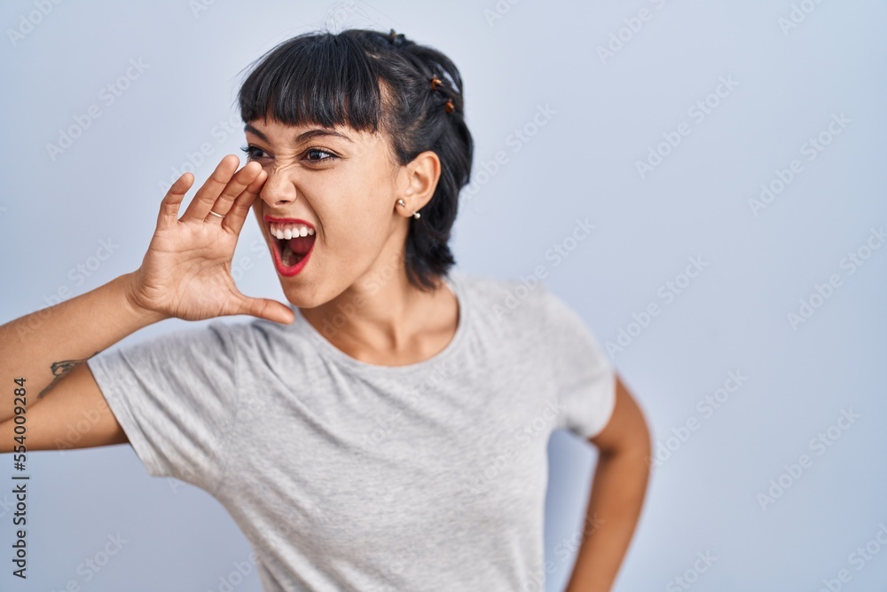 Young hispanic woman wearing casual t shirt over blue background shouting and screaming loud to side with hand on mouth. communication concept. Young hispanic woman wearing casual t shirt over blue background shouting and screaming loud to side with hand on mouth. communication concept.