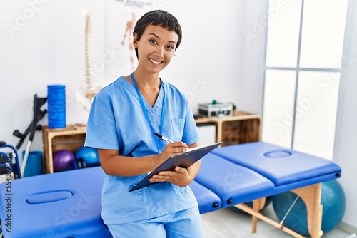 Young hispanic woman wearing physiotherapist uniform writing on checklist at physiotherapy clinic