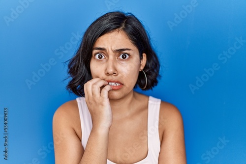 Young hispanic woman standing over blue background looking stressed and nervous with hands on mouth biting nails. anxiety problem.