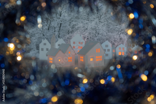 Tableau sur toile Small white cardboard houses with cut shining windows on dark blue bokeh blurred foreground and snowy trees on background