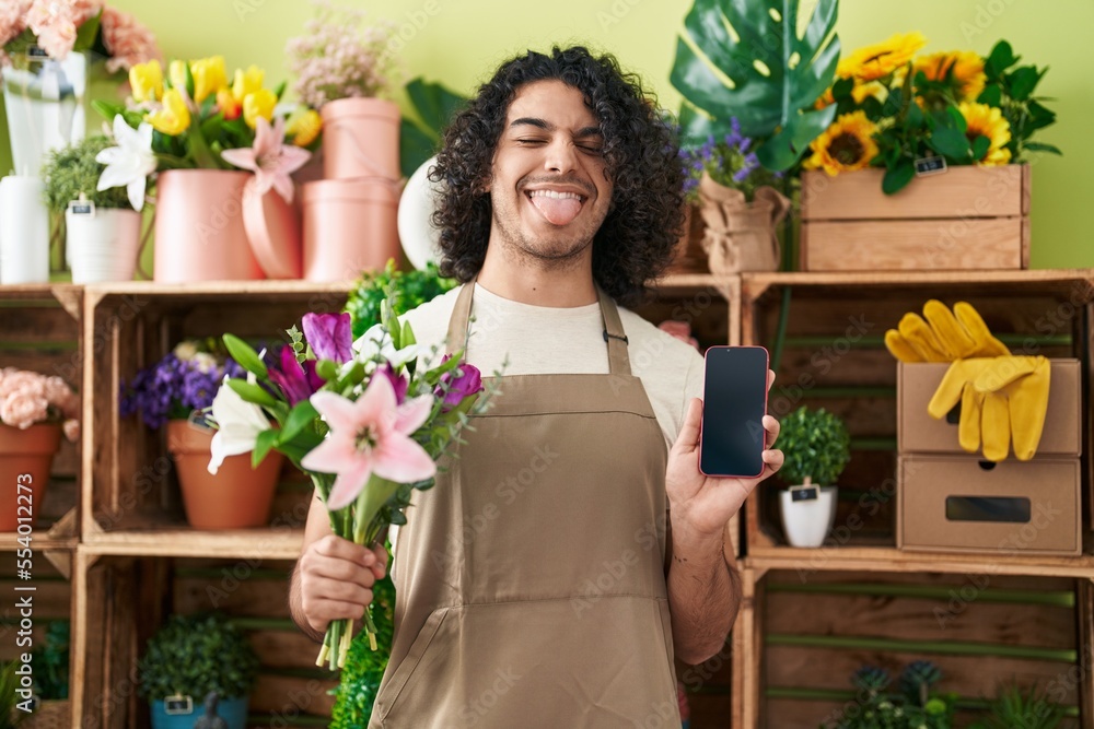 Hispanic man with curly hair working at florist shop showing smartphone screen sticking tongue out happy with funny expression. Hispanic man with curly hair working at florist shop showing smartphone screen sticking tongue out happy with funny expression.