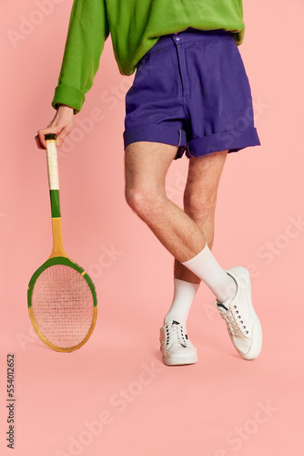 Cropped image of male legs in shorts and sneakers, leaning on tennis racket isolated over pink background. Vintage © master1305