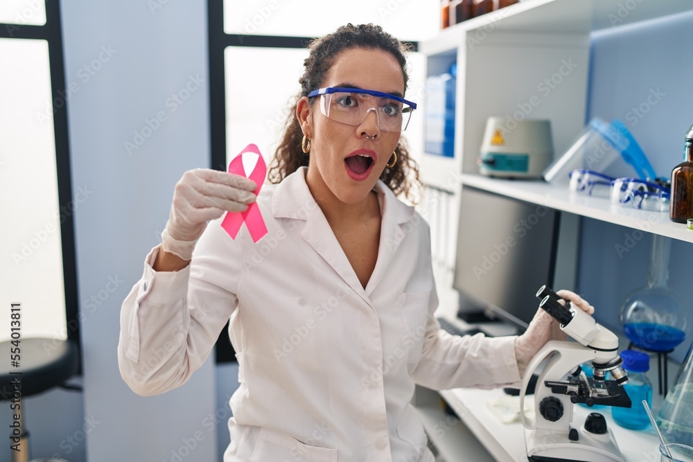 Young hispanic woman working at scientist laboratory looking for breast cancer cure afraid and shocked with surprise and amazed expression, fear and excited face. Young hispanic woman working at scientist laboratory looking for breast cancer cure afraid and shocked with surprise and amazed expression, fear and excited face.