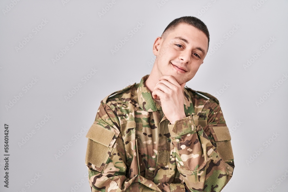 Young man wearing camouflage army uniform looking confident at the camera smiling with crossed arms and hand raised on chin. thinking positive.