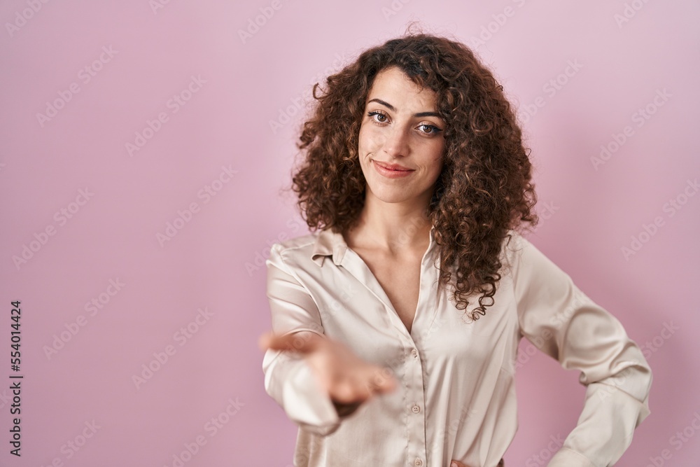 Hispanic woman with curly hair standing over pink background smiling cheerful offering palm hand giving assistance and acceptance. Hispanic woman with curly hair standing over pink background smiling cheerful offering palm hand giving assistance and acceptance.