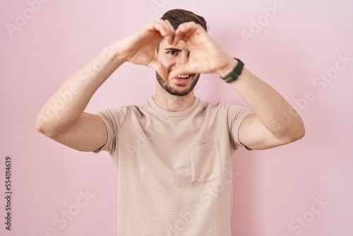 Hispanic man with beard standing over pink background doing heart shape with hand and fingers smiling looking through sign