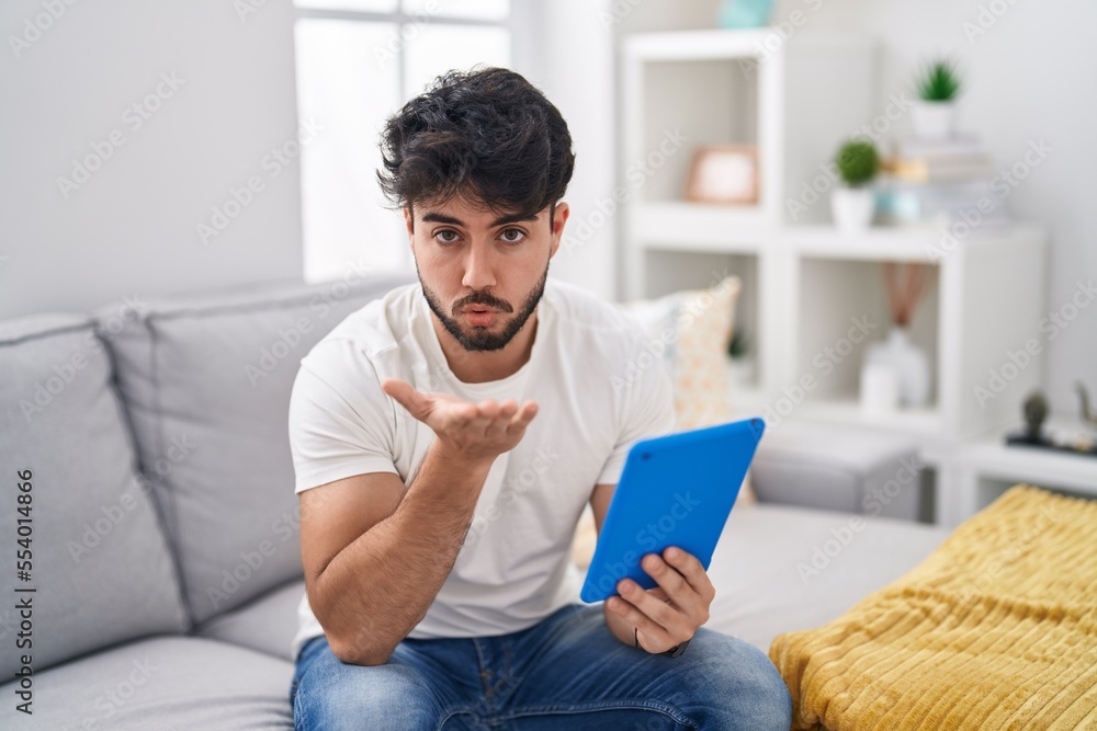Hispanic man with beard using touchpad sitting on the sofa looking at the camera blowing a kiss with hand on air being lovely and sexy. love expression. Hispanic man with beard using touchpad sitting on the sofa looking at the camera blowing a kiss with hand on air being lovely and sexy. love expression.