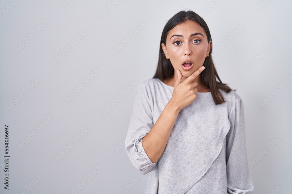 Young hispanic woman standing over white background looking fascinated with disbelief, surprise and amazed expression with hands on chin Young hispanic woman standing over white background looking fascinated with disbelief, surprise and amazed expression with hands on chin