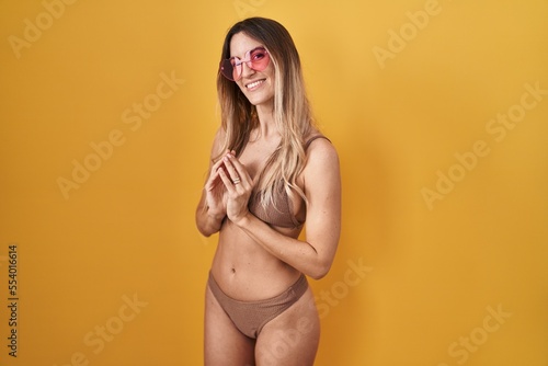 Young hispanic woman wearing bikini over yellow background hands together and fingers crossed smiling relaxed and cheerful. success and optimistic