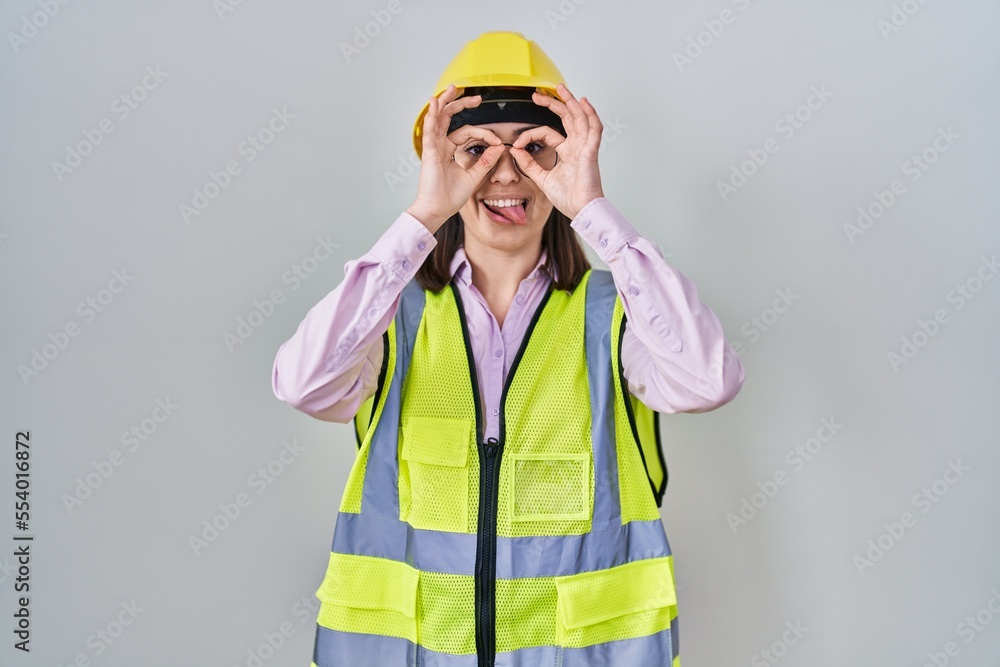 Hispanic girl wearing builder uniform and hardhat doing ok gesture like binoculars sticking tongue out, eyes looking through fingers. crazy expression. Hispanic girl wearing builder uniform and hardhat doing ok gesture like binoculars sticking tongue out, eyes looking through fingers. crazy expression.