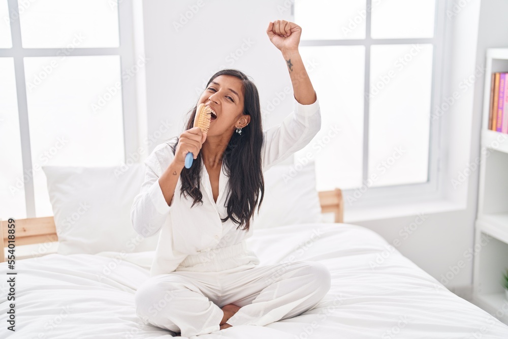 Young beautiful hispanic woman singing song using brush as a microphone at bedroom
