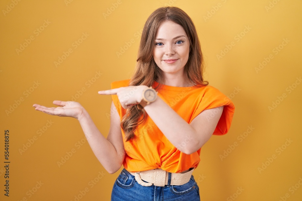 Caucasian woman standing over yellow background amazed and smiling to the camera while presenting with hand and pointing with finger.