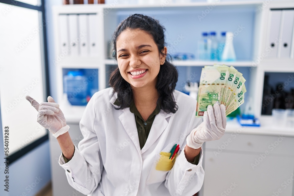 Hispanic woman with dark hair working at scientist laboratory holding money smiling happy pointing with hand and finger to the side Hispanic woman with dark hair working at scientist laboratory holding money smiling happy pointing with hand and finger to the side
