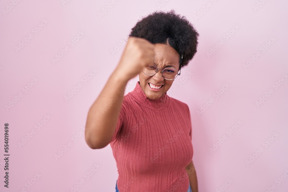 Beautiful african woman with curly hair standing over pink background angry and mad raising fist frustrated and furious while shouting with anger. rage and aggressive concept. Beautiful african woman with curly hair standing over pink background angry and mad raising fist frustrated and furious while shouting with anger. rage and aggressive concept.