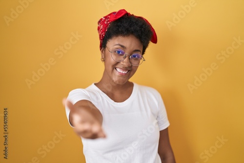 Young african american woman standing over yellow background smiling friendly offering handshake as greeting and welcoming. successful business.