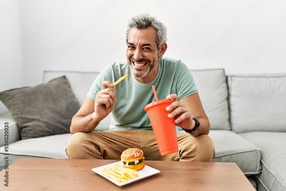 Middle age grey-haired man eating fried potatoes sitting on sofa at home Middle age grey-haired man eating fried potatoes sitting on sofa at home