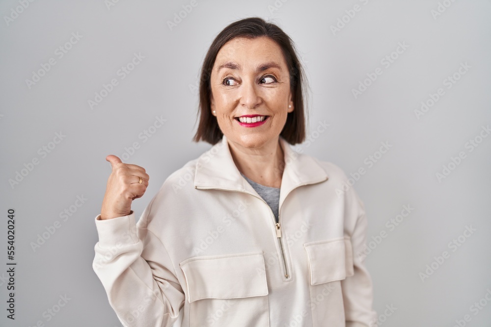 Middle age hispanic woman standing over isolated background smiling with happy face looking and pointing to the side with thumb up. Middle age hispanic woman standing over isolated background smiling with happy face looking and pointing to the side with thumb up.