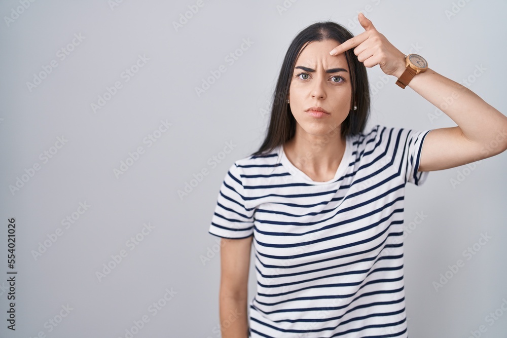 Young brunette woman wearing striped t shirt pointing unhappy to pimple on forehead, ugly infection of blackhead. acne and skin problem Young brunette woman wearing striped t shirt pointing unhappy to pimple on forehead, ugly infection of blackhead. acne and skin problem