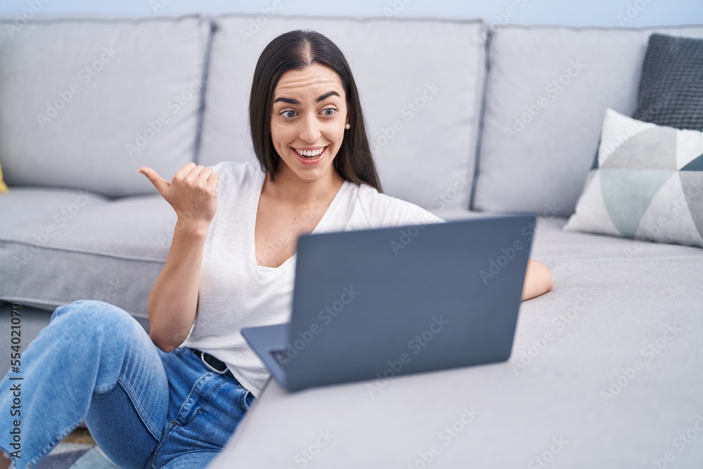 Young brunette woman using laptop at home pointing thumb up to the side smiling happy with open mouth Young brunette woman using laptop at home pointing thumb up to the side smiling happy with open mouth