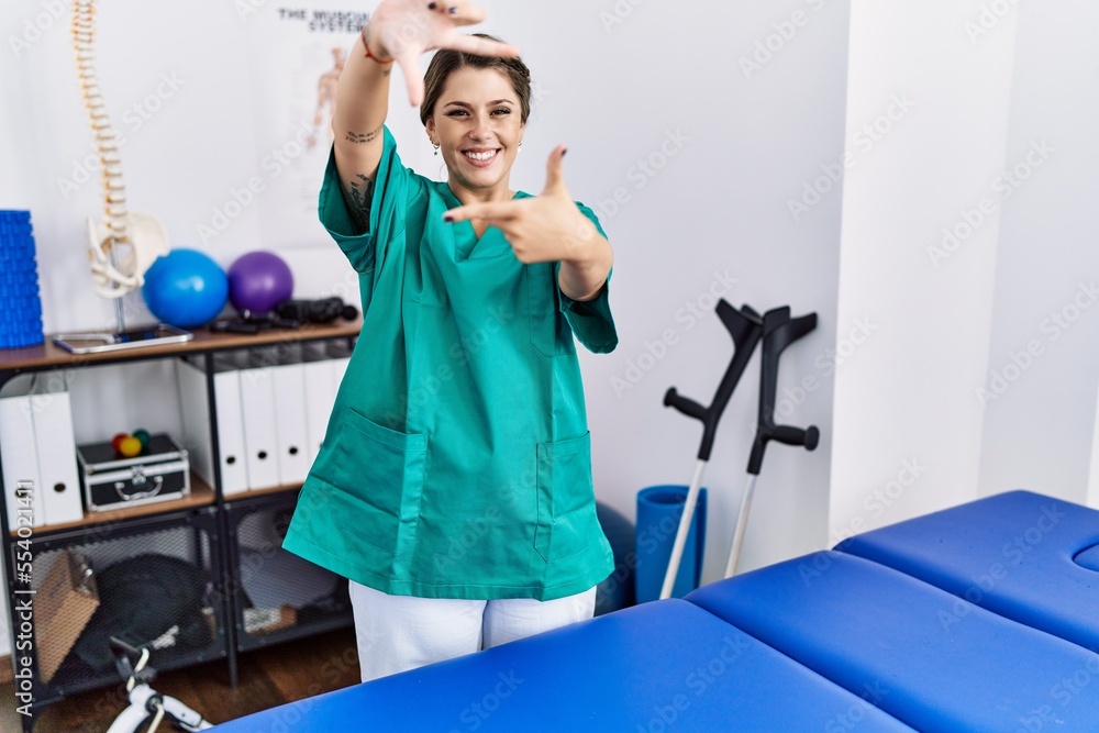 Young hispanic woman wearing physiotherapist uniform standing at clinic smiling making frame with hands and fingers with happy face. creativity and photography concept. Young hispanic woman wearing physiotherapist uniform standing at clinic smiling making frame with hands and fingers with happy face. creativity and photography concept.
