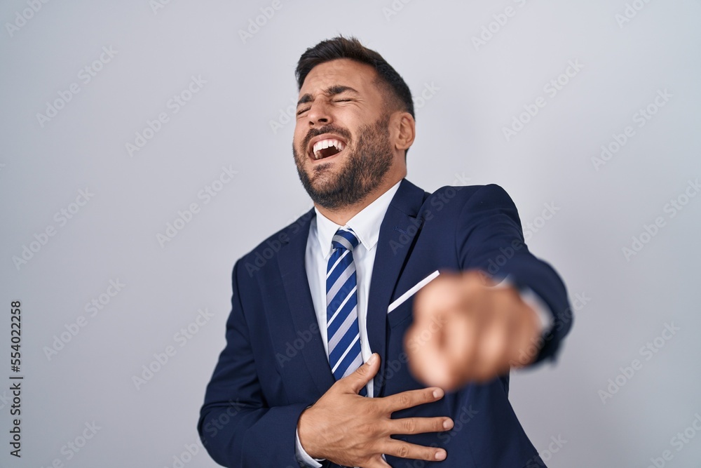 Handsome hispanic man wearing suit and tie laughing at you, pointing finger to the camera with hand over body, shame expression Handsome hispanic man wearing suit and tie laughing at you, pointing finger to the camera with hand over body, shame expression