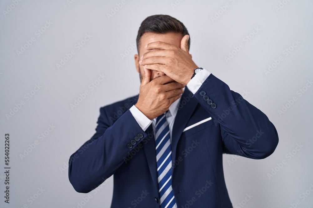 Handsome hispanic man wearing suit and tie covering eyes and mouth with hands, surprised and shocked. hiding emotion Handsome hispanic man wearing suit and tie covering eyes and mouth with hands, surprised and shocked. hiding emotion