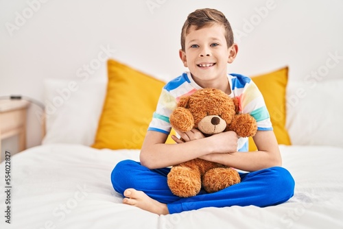 Blond child hugging teddy bear sitting on bed at bedroom