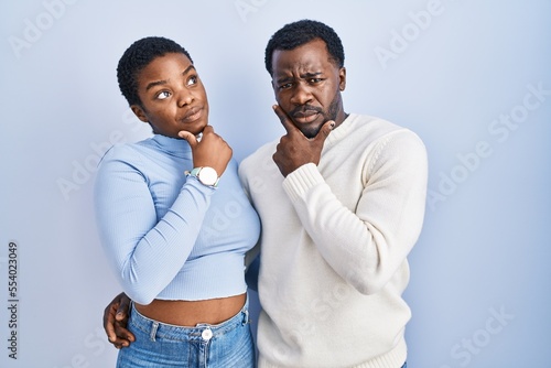 Young african american couple standing over blue background looking confident at the camera smiling with crossed arms and hand raised on chin. thinking positive.