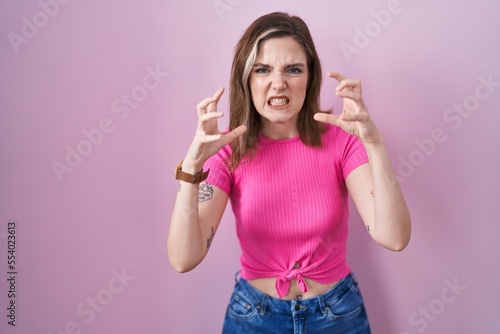 Blonde caucasian woman standing over pink background shouting frustrated with rage  hands trying to strangle  yelling mad