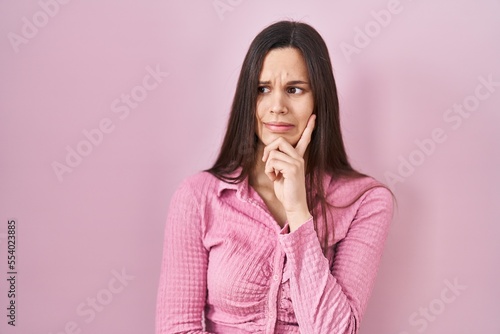 Young hispanic woman standing over pink background thinking worried about a question, concerned and nervous with hand on chin