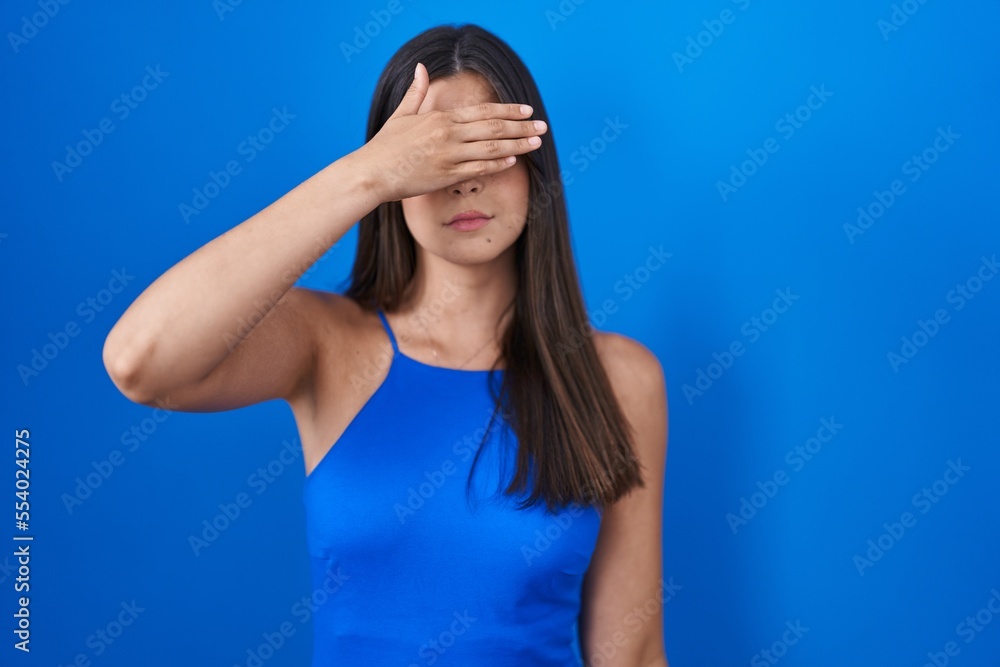Hispanic woman standing over blue background covering eyes with hand, looking serious and sad. sightless, hiding and rejection concept Hispanic woman standing over blue background covering eyes with hand, looking serious and sad. sightless, hiding and rejection concept