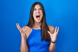 Hispanic woman standing over blue background crazy and mad shouting and yelling with aggressive expression and arms raised. frustration concept.