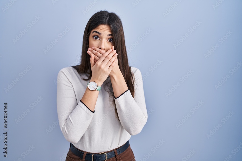 Young brunette woman standing over blue background shocked covering mouth with hands for mistake. secret concept. Young brunette woman standing over blue background shocked covering mouth with hands for mistake. secret concept.