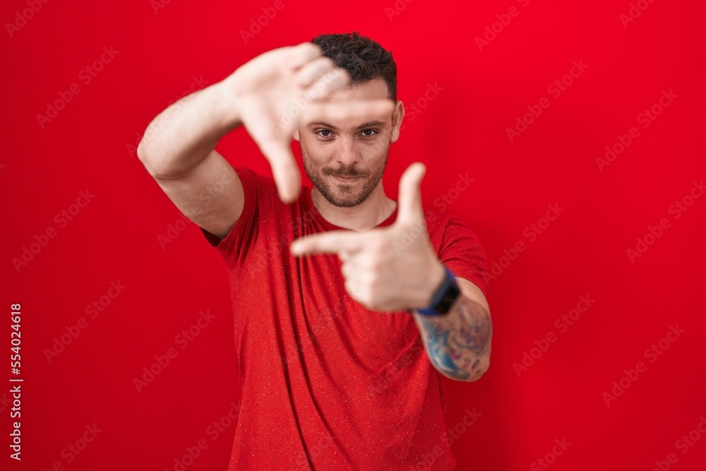Young hispanic man standing over red background smiling making frame with hands and fingers with happy face. creativity and photography concept. Young hispanic man standing over red background smiling making frame with hands and fingers with happy face. creativity and photography concept.