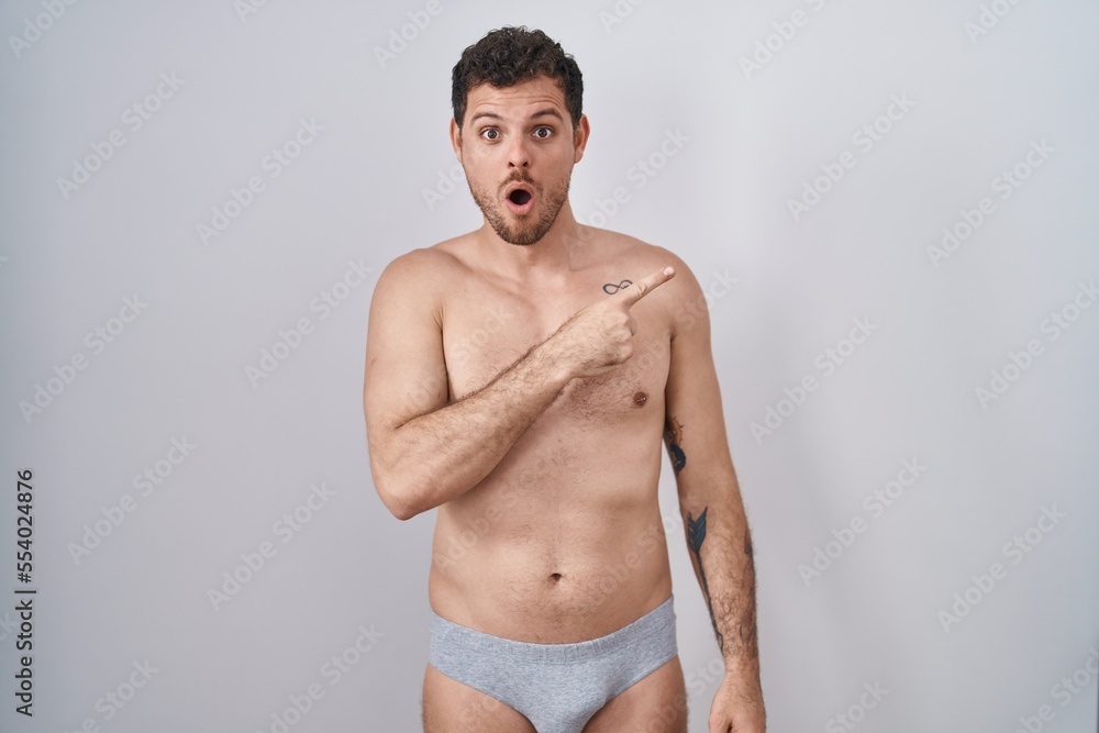Young hispanic man standing shirtless wearing underware surprised pointing with finger to the side, open mouth amazed expression.