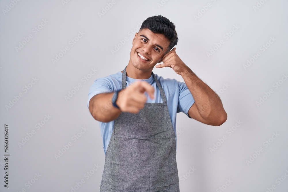 Hispanic young man wearing apron over white background smiling doing talking on the telephone gesture and pointing to you. call me. Hispanic young man wearing apron over white background smiling doing talking on the telephone gesture and pointing to you. call me.