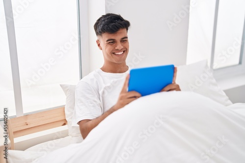 Young hispanic man using touchpad sitting on bed at bedroom