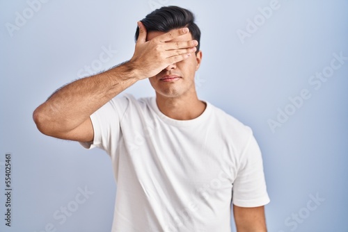 Картината върху платно Hispanic man standing over blue background covering eyes with hand, looking serious and sad