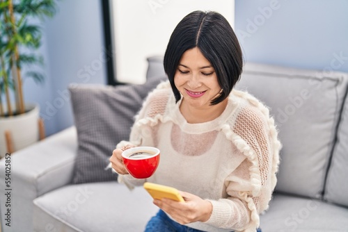 Young chinese woman drinking coffee and using smartphone at home