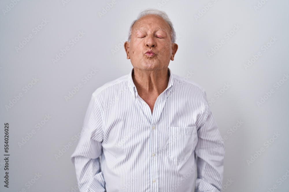 Senior man with grey hair standing over isolated background looking at the camera blowing a kiss on air being lovely and sexy. love expression. Senior man with grey hair standing over isolated background looking at the camera blowing a kiss on air being lovely and sexy. love expression.