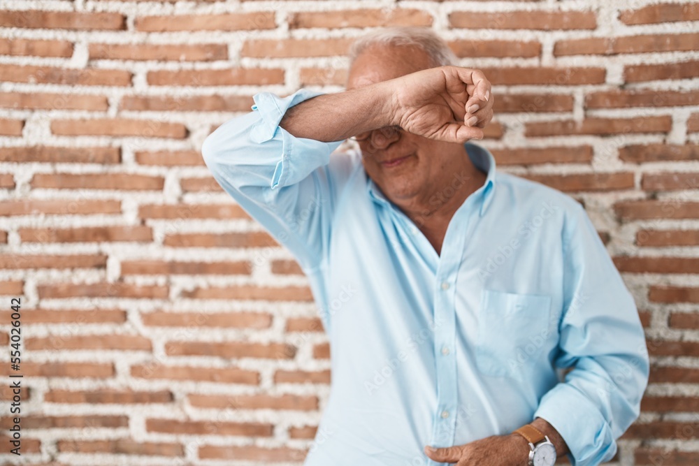 Senior man with grey hair standing over bricks wall covering eyes with arm smiling cheerful and funny. blind concept. Senior man with grey hair standing over bricks wall covering eyes with arm smiling cheerful and funny. blind concept.