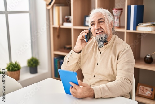 Middle age grey-haired man talking on smartphone using touchpad at home