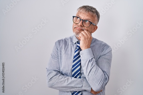 Hispanic business man with grey hair wearing glasses with hand on chin thinking about question, pensive expression. smiling with thoughtful face. doubt concept.